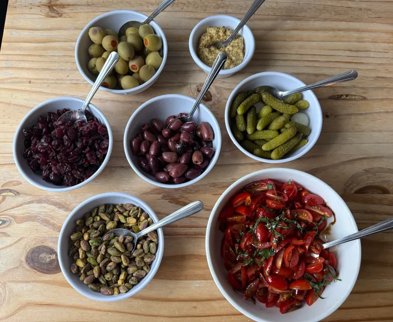 Bowls of salad-bar toppings — green olives, dried cranberries, kalamata olives, cornichons, pistachios, and marinated tomatoes