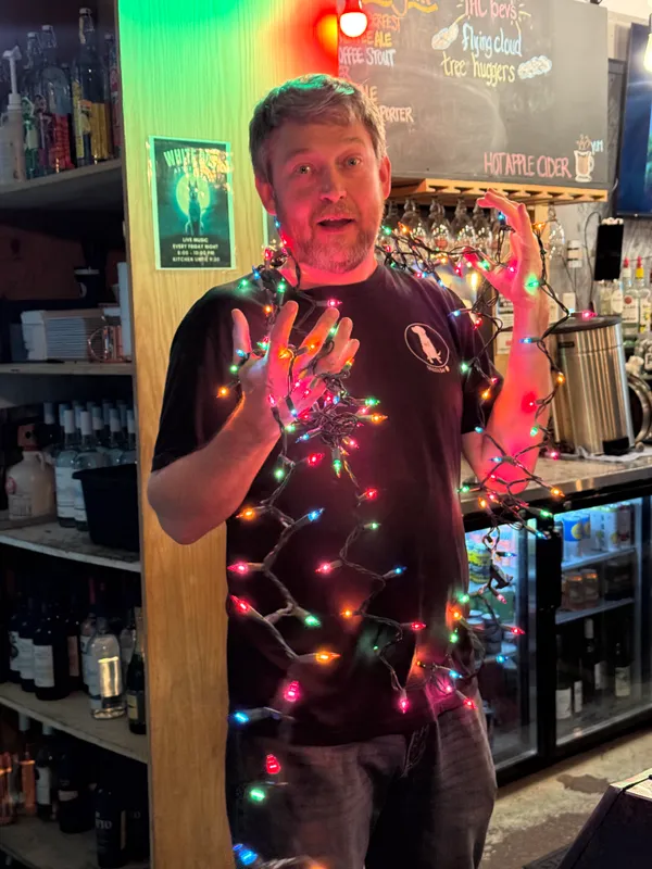 A staff member playfully wrapped in colorful Christmas lights behind the bar in a White Dog tee