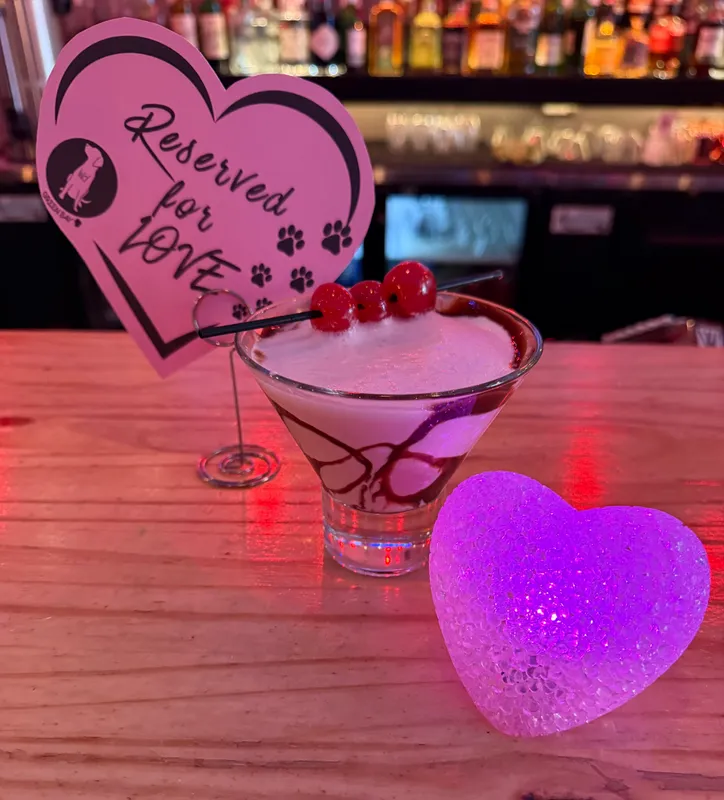 A creamy Valentine's cocktail garnished with cherries beside a glowing pink heart and a "Reserved for Love" sign on the bar